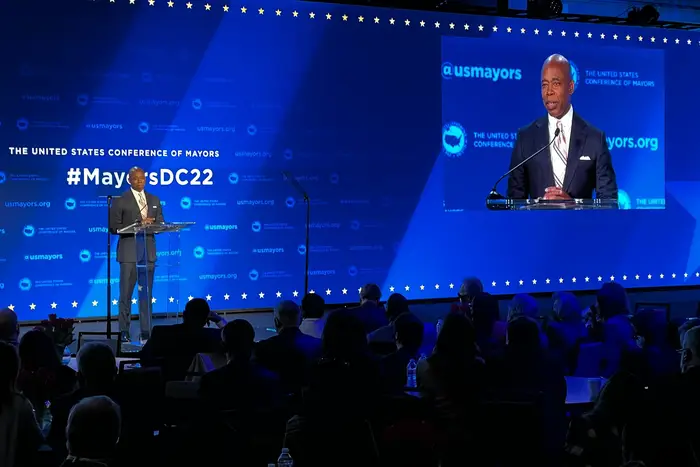 Mayor Eric Adams, standing on a stage with a blue background as a big screen TV hung from the ceiling broadcasts a video of him, offers remarks at the United States Conference of Mayors in Washington, D.C. on January 20th.